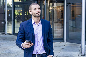 Ein Mann in einem blauen Anzug mit Smartphone in der Hand posiert vor einem modernen Bürogebäude für ein Business-Fotoshooting.