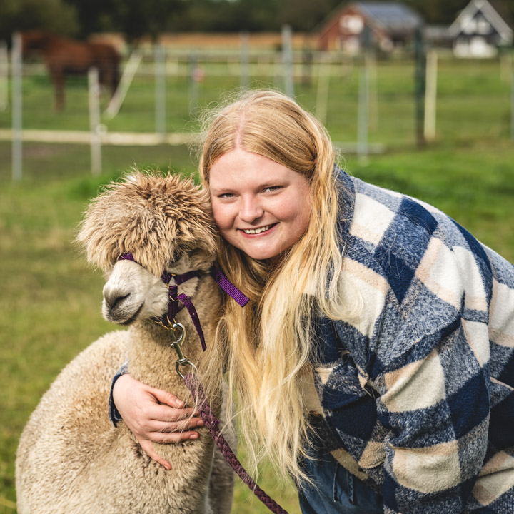 Ein professionelles Porträt einer jungen, blonden Frau, die ein Alpaka umarmt.
